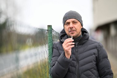 Kışlık ceketli ve bereli bir adam dışarıdaki tel örgülerin yanında sigara içiyor..