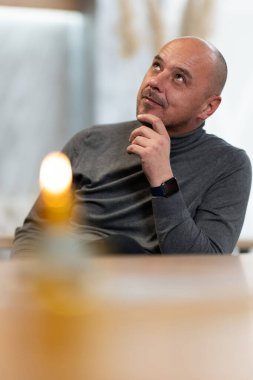 Eli çenesinde düşünen kel bir adam yukarı bakıyor, içeride oturuyor..