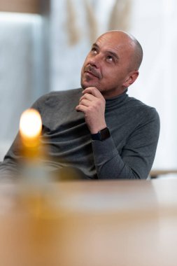 Eli çenesinde düşünen kel bir adam yukarı bakıyor, içeride oturuyor..