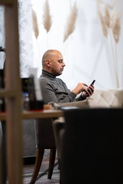 Gri balıkçı yaka giyen kel adam evde otururken akıllı telefonunu kullanıyor..