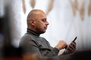 Gri balıkçı yaka giyen kel adam evde otururken akıllı telefonunu kullanıyor..