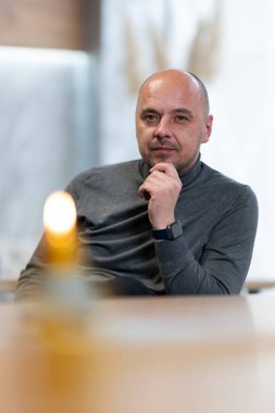 Boğazlı kazaklı, sırıtan kel bir adam. Önünde mum olan bir masada oturuyor..