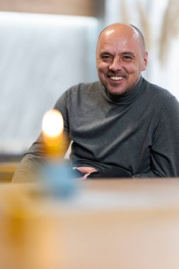 Boğazlı kazaklı, sırıtan kel bir adam. Önünde mum olan bir masada oturuyor..