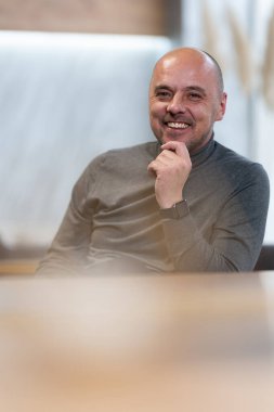 Boğazlı kazaklı, sırıtan kel bir adam. Önünde mum olan bir masada oturuyor..