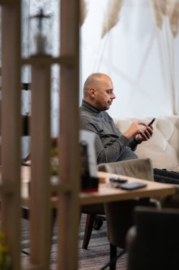 Gri balıkçı yaka giyen kel adam evde otururken akıllı telefonunu kullanıyor..