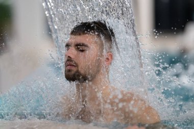 Spa 'da hidroterapi şelalesi. Bir adam kaplıca havuzunda hidroterapi şelalesinin altında duruyor, kafasına ve omuzlarına su masajı yaptırıyor..