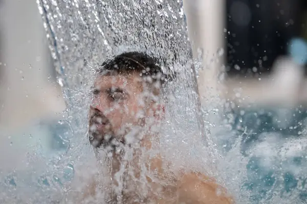 Spa 'da hidroterapi şelalesi. Bir adam kaplıca havuzunda hidroterapi şelalesinin altında duruyor, kafasına ve omuzlarına su masajı yaptırıyor..