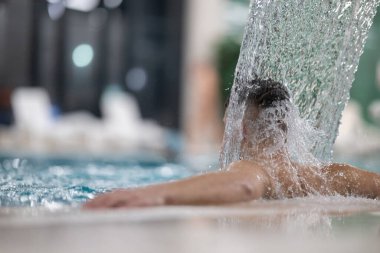 Spa 'da hidroterapi şelalesi. Bir adam kaplıca havuzunda hidroterapi şelalesinin altında duruyor, kafasına ve omuzlarına su masajı yaptırıyor..