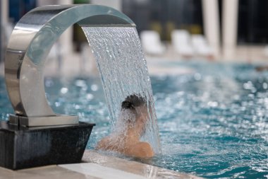 Spa 'da hidroterapi şelalesi. Bir adam kaplıca havuzunda hidroterapi şelalesinin altında duruyor, kafasına ve omuzlarına su masajı yaptırıyor..