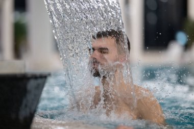 Spa 'da hidroterapi şelalesi. Bir adam kaplıca havuzunda hidroterapi şelalesinin altında duruyor, kafasına ve omuzlarına su masajı yaptırıyor..