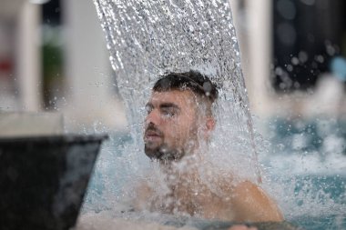 Spa 'da hidroterapi şelalesi. Bir adam kaplıca havuzunda hidroterapi şelalesinin altında duruyor, kafasına ve omuzlarına su masajı yaptırıyor..