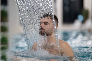 Spa 'da hidroterapi şelalesi. Bir adam kaplıca havuzunda hidroterapi şelalesinin altında duruyor, kafasına ve omuzlarına su masajı yaptırıyor..