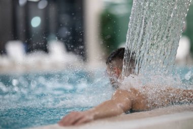 Spa 'da hidroterapi şelalesi. Bir adam kaplıca havuzunda hidroterapi şelalesinin altında duruyor, kafasına ve omuzlarına su masajı yaptırıyor..
