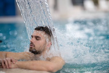 Spa 'da hidroterapi şelalesi. Bir adam kaplıca havuzunda hidroterapi şelalesinin altında duruyor, kafasına ve omuzlarına su masajı yaptırıyor..