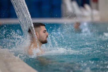 Spa 'da hidroterapi şelalesi. Bir adam kaplıca havuzunda hidroterapi şelalesinin altında duruyor, kafasına ve omuzlarına su masajı yaptırıyor..
