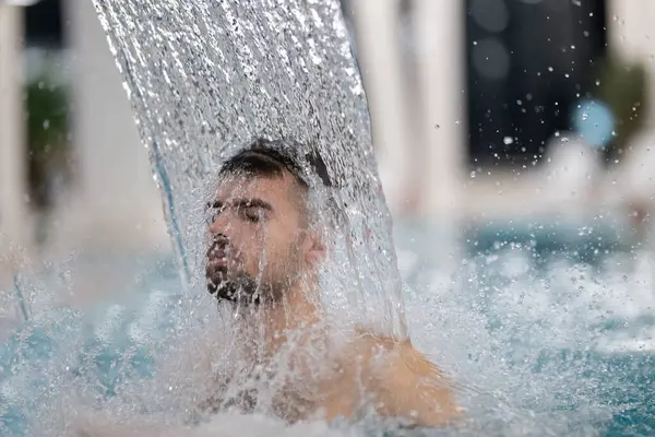 Spa 'da hidroterapi şelalesi. Bir adam kaplıca havuzunda hidroterapi şelalesinin altında duruyor, kafasına ve omuzlarına su masajı yaptırıyor..