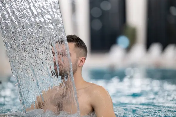 Spa 'da hidroterapi şelalesi. Bir adam kaplıca havuzunda hidroterapi şelalesinin altında duruyor, kafasına ve omuzlarına su masajı yaptırıyor..