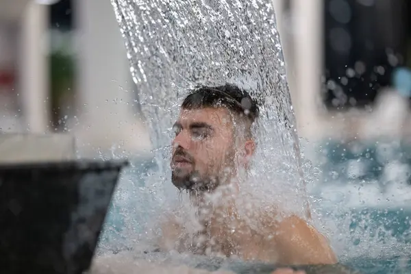 Spa 'da hidroterapi şelalesi. Bir adam kaplıca havuzunda hidroterapi şelalesinin altında duruyor, kafasına ve omuzlarına su masajı yaptırıyor..