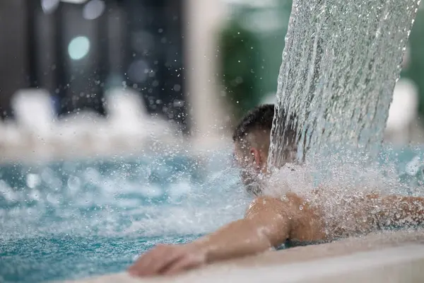 Spa 'da hidroterapi şelalesi. Bir adam kaplıca havuzunda hidroterapi şelalesinin altında duruyor, kafasına ve omuzlarına su masajı yaptırıyor..