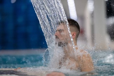 Spa 'da hidroterapi şelalesi. Bir adam kaplıca havuzunda hidroterapi şelalesinin altında duruyor, kafasına ve omuzlarına su masajı yaptırıyor..