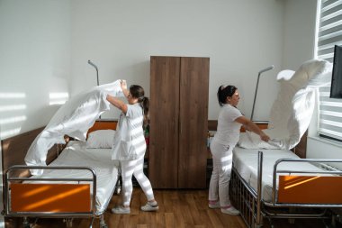 Üniformalı iki hemşire çarşafları değiştirerek ve hastalar için hijyenik bir ortam sağlayarak hastane odasını temizleyip düzenliyor.