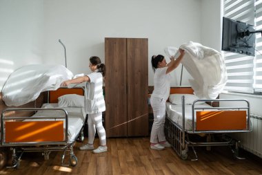 Üniformalı iki hemşire çarşafları değiştirerek ve hastalar için hijyenik bir ortam sağlayarak hastane odasını temizleyip düzenliyor.