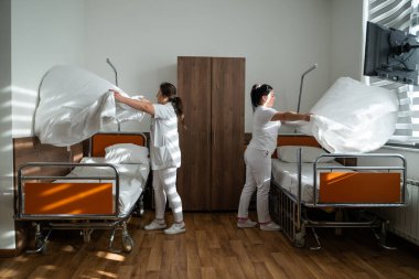 Üniformalı iki hemşire çarşafları değiştirerek ve hastalar için hijyenik bir ortam sağlayarak hastane odasını temizleyip düzenliyor.