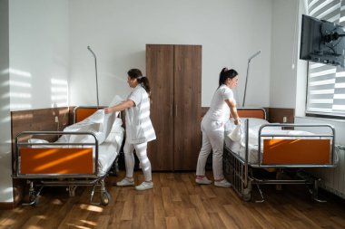 Üniformalı iki hemşire çarşafları değiştirerek ve hastalar için hijyenik bir ortam sağlayarak hastane odasını temizleyip düzenliyor.