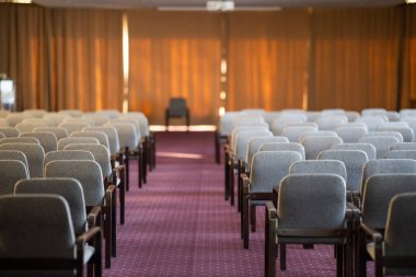 Geniş bir konferans salonu. Sıralar halinde gri sandalyeler ön tarafta tek sandalyeye bakıyor. Oda, büyük perdelerin arasından süzülen sıcak doğal ışıkla aydınlanıyor.