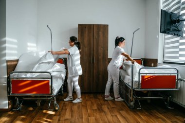 Üniformalı iki hemşire çarşafları değiştirerek ve hastalar için hijyenik bir ortam sağlayarak hastane odasını temizleyip düzenliyor.
