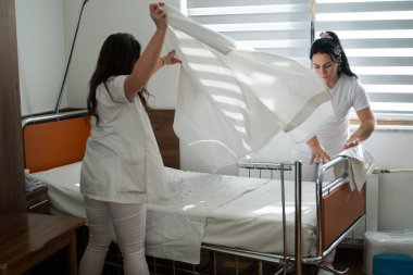 Üniformalı iki hemşire çarşafları değiştirerek ve hastalar için hijyenik bir ortam sağlayarak hastane odasını temizleyip düzenliyor.