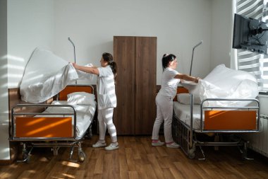 Üniformalı iki hemşire çarşafları değiştirerek ve hastalar için hijyenik bir ortam sağlayarak hastane odasını temizleyip düzenliyor.