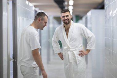 Spa Koridoru 'nda tıbbi danışmanlık. İki adam klinik spa koridorunda yürürken tedavi seçeneklerini tartışıyor, beyaz üniforma ve bornoz giyiyor.