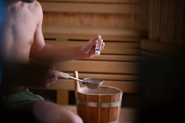 Geleneksel ahşap bir saunanın içinde elinde kova ve kepçeyle gerekli yağları tutan bir adamın eli yakındır..