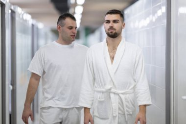 Spa Koridoru 'nda tıbbi danışmanlık. İki adam klinik spa koridorunda yürürken tedavi seçeneklerini tartışıyor, beyaz üniforma ve bornoz giyiyor.