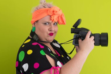 Parlak yeşil arka planda, canlı kıyafetli, profesyonel kamerayla çekim yapan iri yarı bir kadının yan portresi.. 