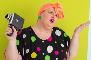 Artı renkli polka noktalı elbiseli kadın elinde retro kamera ve yeşil arkaplan önünde kuşkulu ve etkilenmemiş bir ifade yapıyor.. 