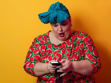 Artı deniz mavisi bir başlık ve karpuz elbise giyen bir kadın canlı turuncu bir arka planda akıllı telefonuna şaşırmış bir ifadeyle bakıyor..