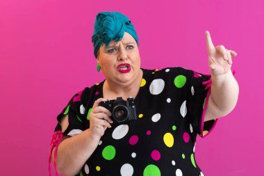 Kendine güvenen, puantiyeli elbiseli, elinde fotoğraf makinesi olan, pembe stüdyo arka planında izole edilmiş bir kadın.. 