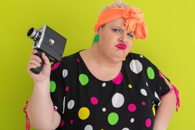 Artı renkli polka noktalı elbiseli kadın elinde retro kamera ve yeşil arkaplan önünde kuşkulu ve etkilenmemiş bir ifade yapıyor.. 