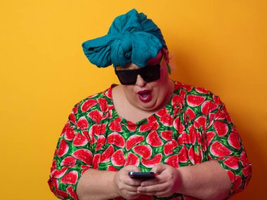 Artı deniz mavisi bir başlık ve karpuz elbise giyen bir kadın canlı turuncu bir arka planda akıllı telefonuna şaşırmış bir ifadeyle bakıyor..