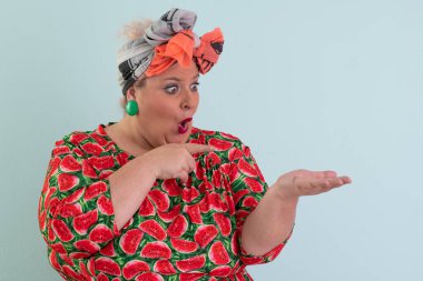 Karpuz desenli elbisesiyle açık avucuna komik ve şaşkın bakışlı açık mavi arkaplana bakan etkileyici ve iri yarı kadın.. 