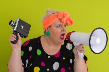 Turuncu başlıklı, etkileyici, iri yarı kadın yeşil arka planda elinde eski model bir kamerayla megafona bağırıyor.. 