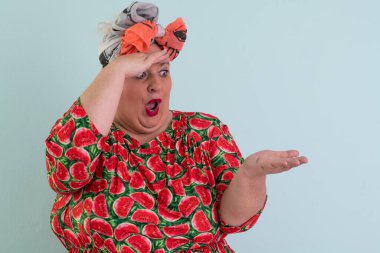 Karpuz desenli elbisesiyle açık avucuna komik ve şaşkın bakışlı açık mavi arkaplana bakan etkileyici ve iri yarı kadın.. 