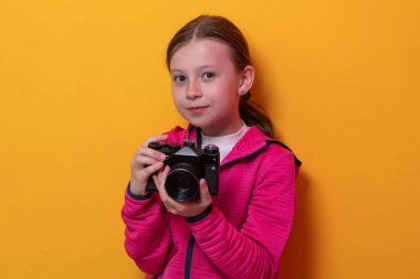 Pembe kapüşonlu şirin bir kız elinde eski model bir kamera tutarken parlak turuncu stüdyo arka planına karşı duruyor.. 