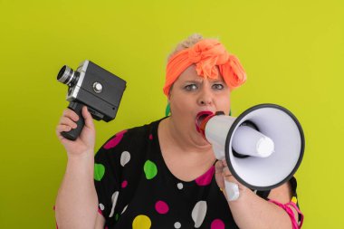 Turuncu başlıklı, etkileyici, iri yarı kadın yeşil arka planda elinde eski model bir kamerayla megafona bağırıyor.. 