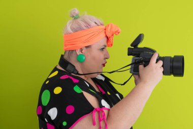 Parlak yeşil arka planda, canlı kıyafetli, profesyonel kamerayla çekim yapan iri yarı bir kadının yan portresi.. 