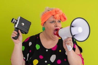 Turuncu başlıklı, etkileyici, iri yarı kadın yeşil arka planda elinde eski model bir kamerayla megafona bağırıyor.. 