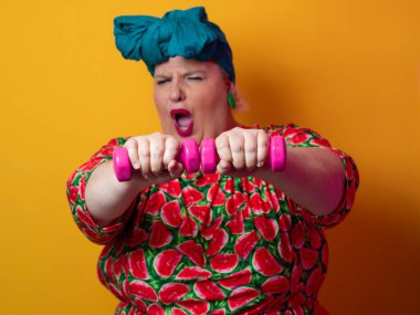 Artı renkli karpuz desenli elbise ve deniz mavisi başlıklı kadın turuncu bir arka plana karşı komik ve konsantre bir yüz ifadesiyle pembe halterleri kaldırıyor..