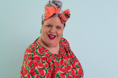 Plus size woman in a colorful watermelon themed dress and orange headwrap posing with unimpressed facial expression and hands on hips against pastel blue background.  clipart